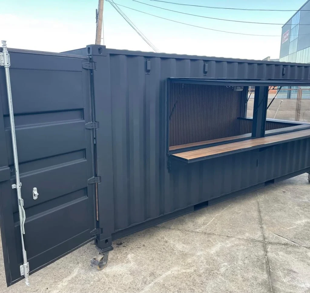 Black shipping container converted into a bar with a fold-up serving window and wood counter