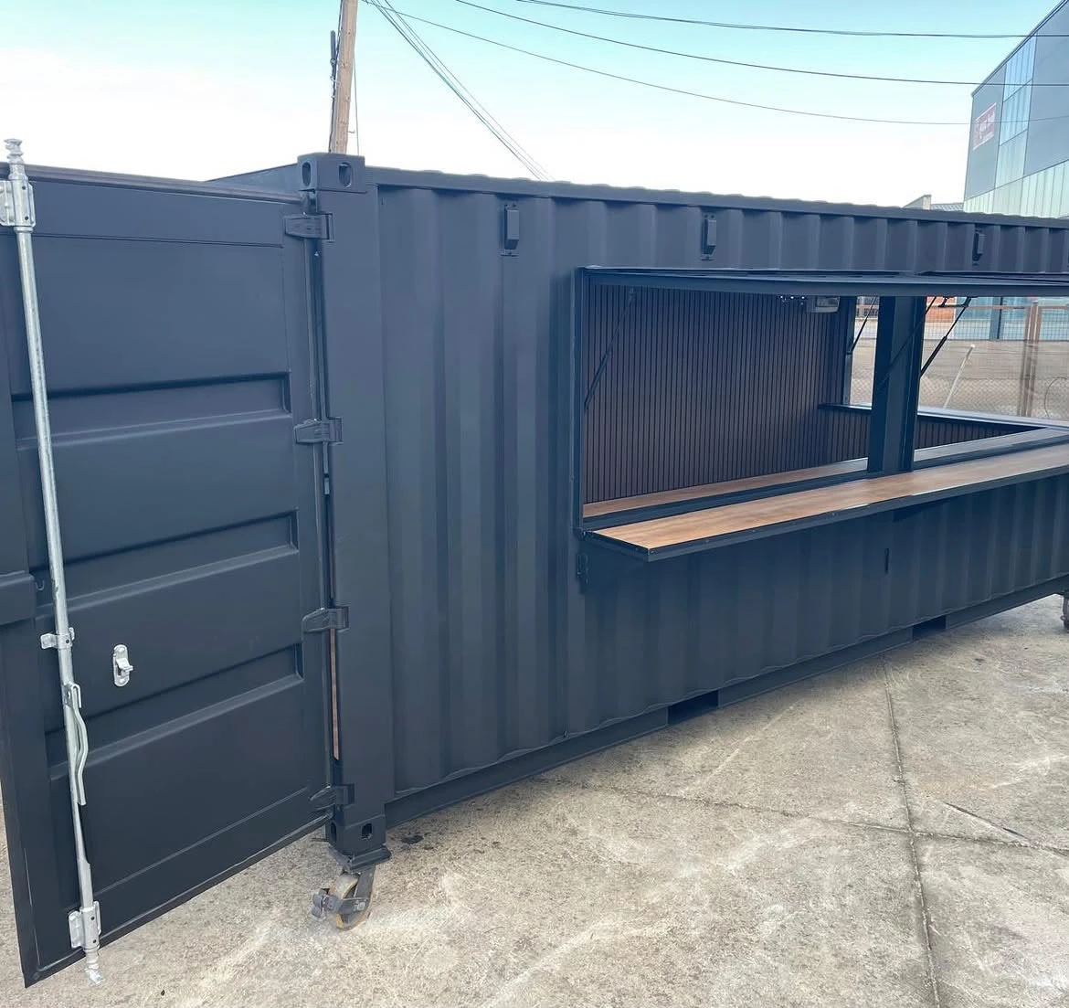 Black shipping container converted into a bar with a fold-up serving window and wood counter